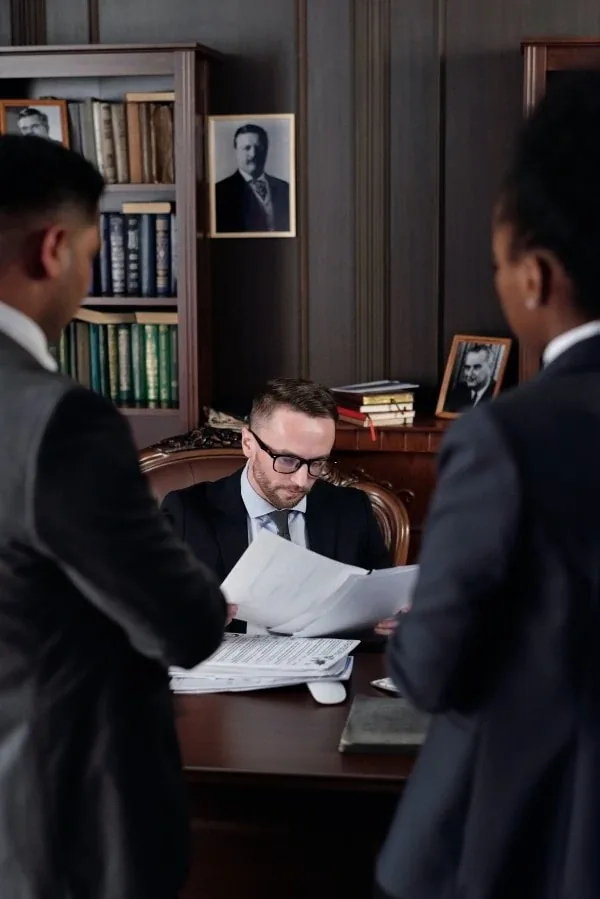 A man in a suit reading paperwork at a desk while two people look on